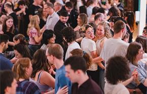 A large, joyful crowd of people dance closely together in a warmly lit room. Smiling faces and casual attire create a lively, festive atmosphere.